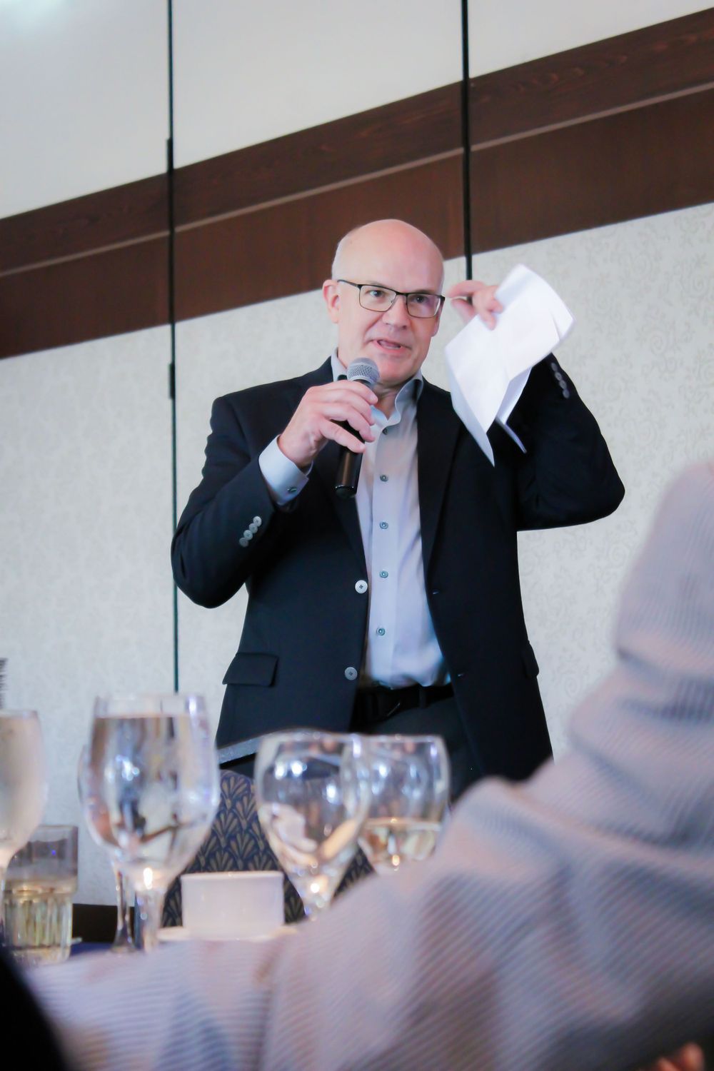 Speaker in a suit gestures while giving a speech at a formal event with wine glasses on tables.