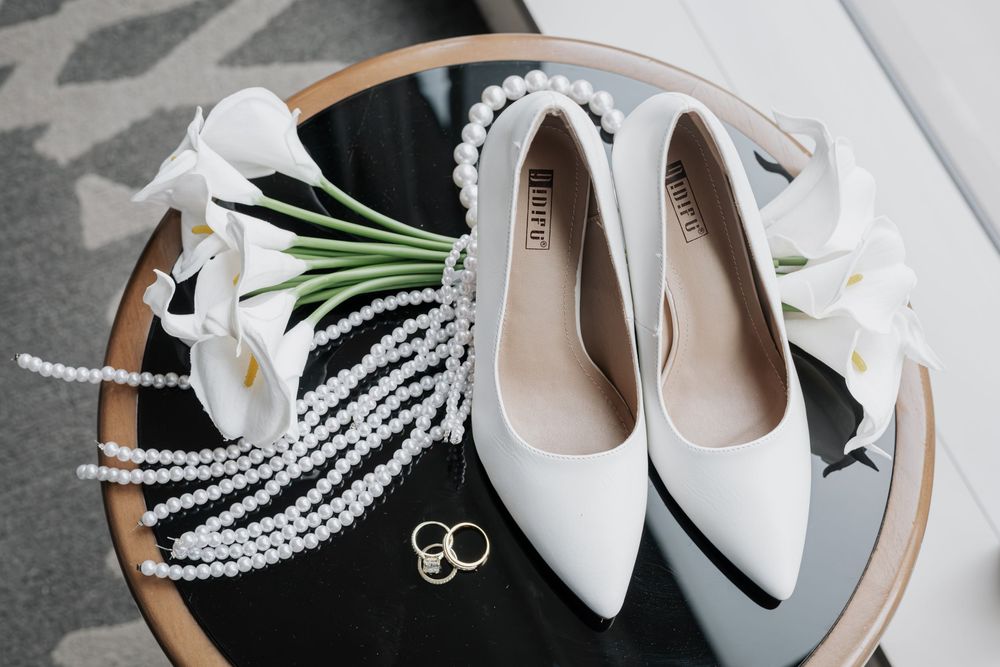 White bridal heels with pearl necklaces, gold rings, and white calla lilies arranged on a round glass table.