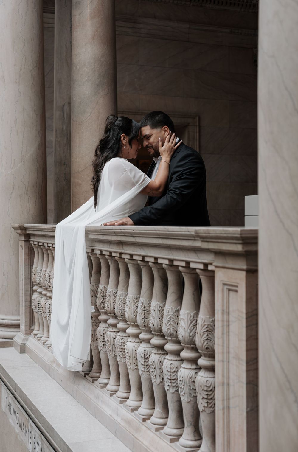 Couple sharing a romantic kiss on an ornate stone balcony with classical columns, bride in white gown.