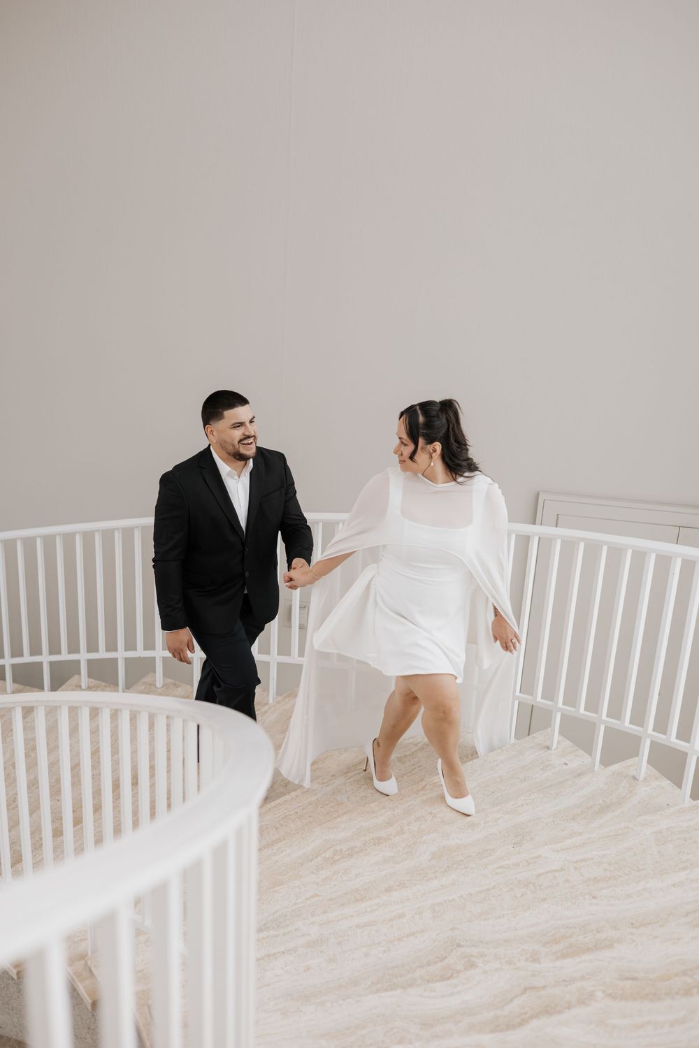 A couple in wedding attire holds hands while walking up a white spiral staircase in a bright, elegant venue.