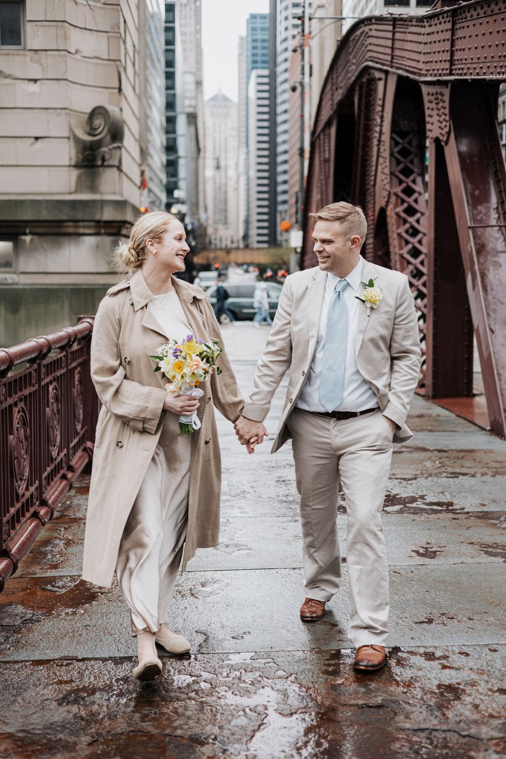 Newlywed couple holding hands walking on a rainy Chicago bridge, bride in trench coat with yellow bouquet.