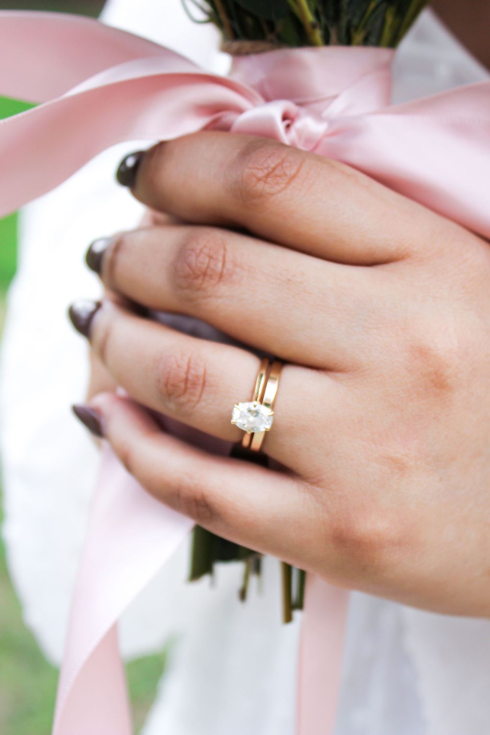 Close up of an engagement ring and wedding band on hand holding flowers with pink ribbon.