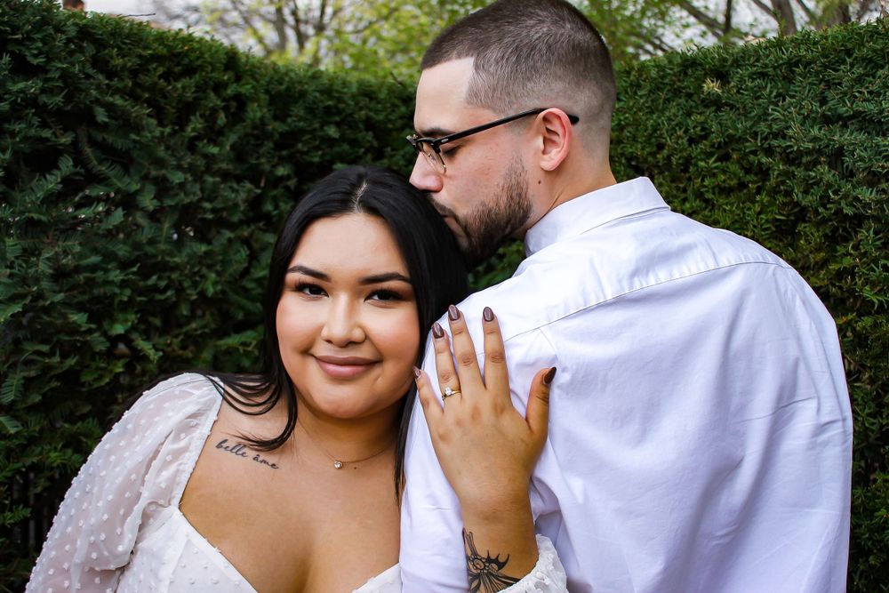 Engaged couple posing together in front of green hedge.
