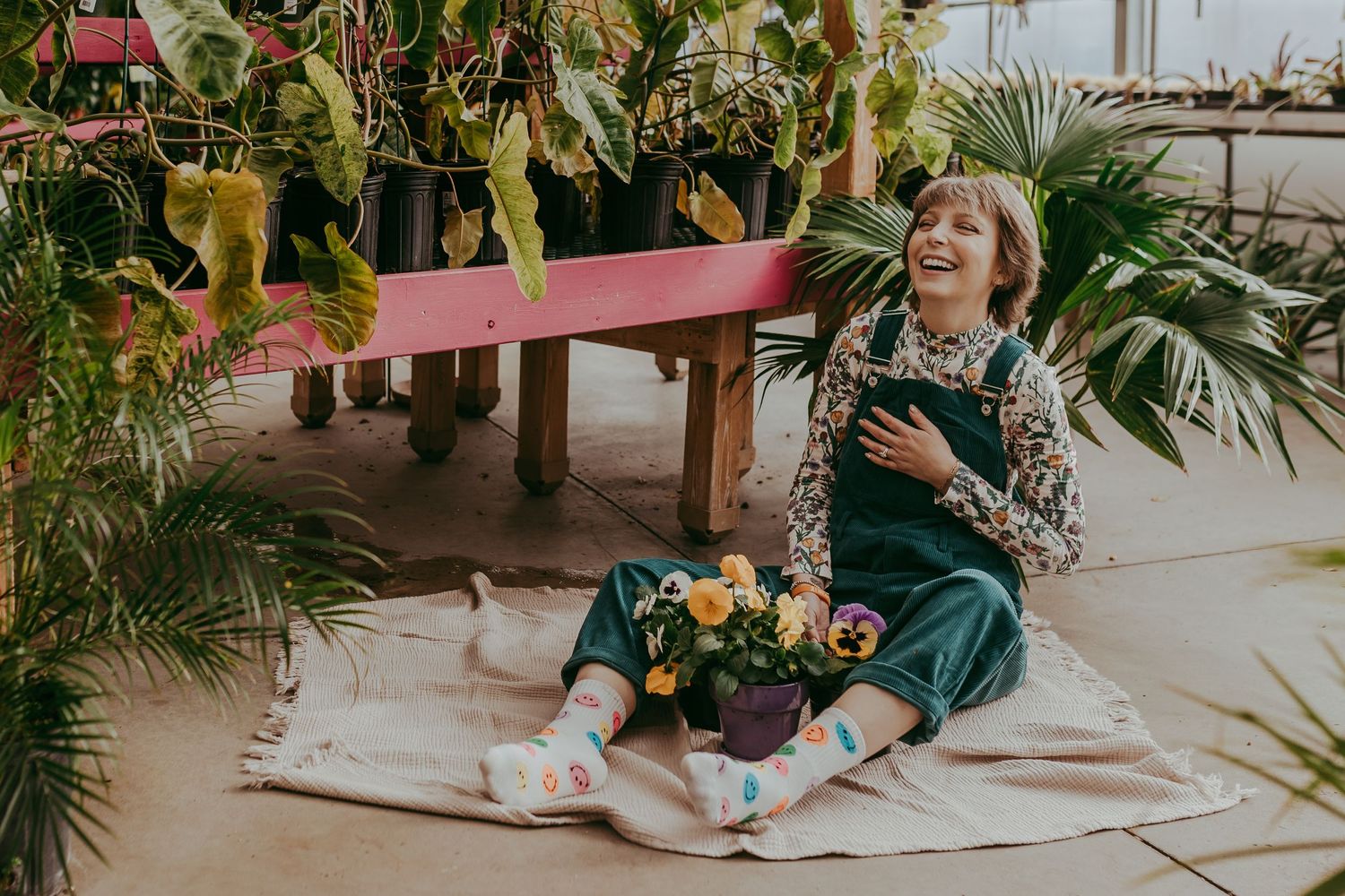 Person in green overalls sits on blanket surrounded by lush plants and flowers in greenhouse setting.