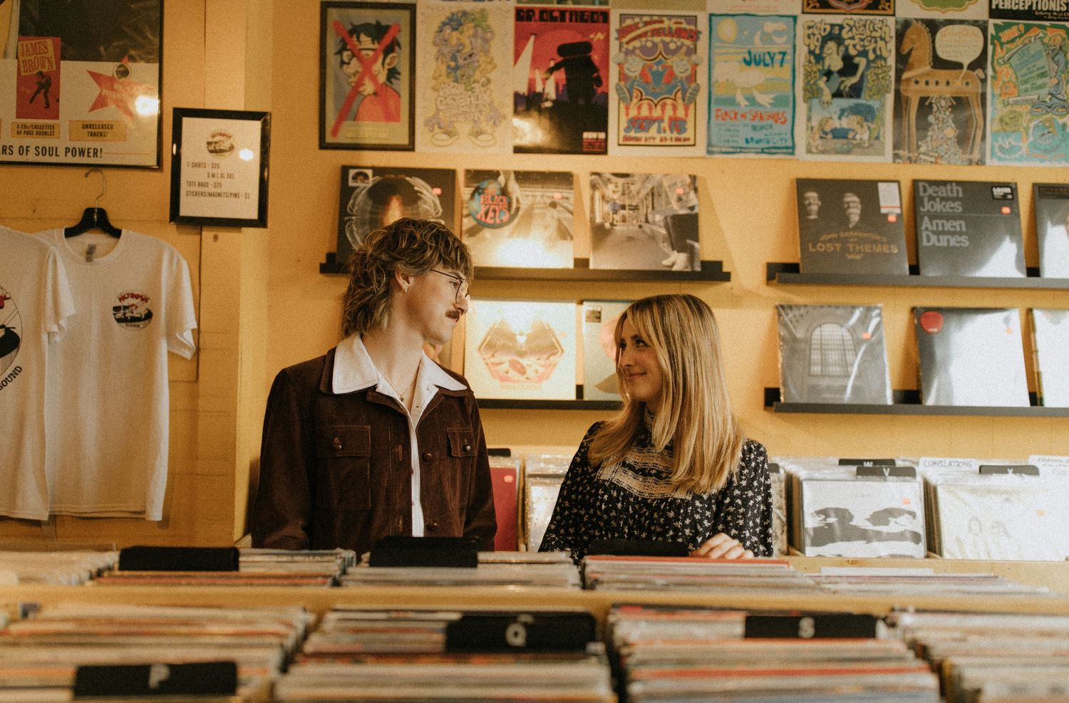 Two individuals browse through records at a vintage record store with posters on the wall.