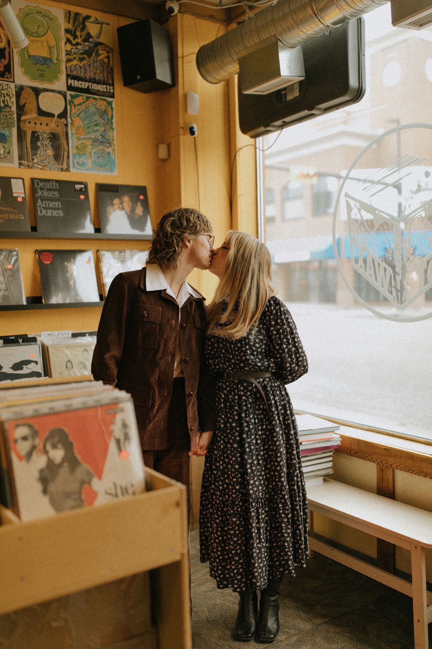 A couple shares an intimate moment while browsing vinyl records in a cozy record store with posters on yellow walls.