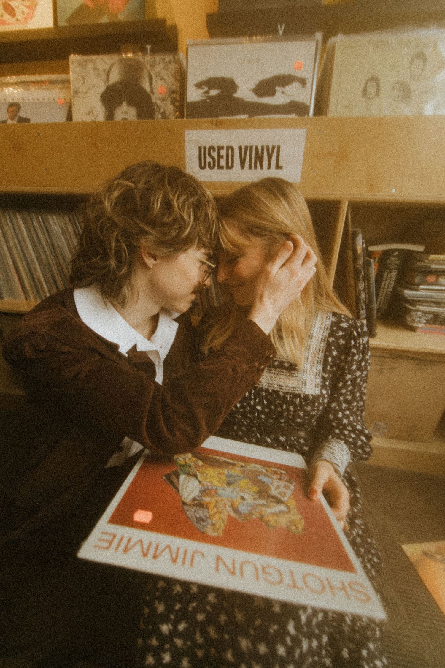 A vintage-tinted photo of two people browsing vinyl records together in a record store, with a 'Used Vinyl' sign visible.