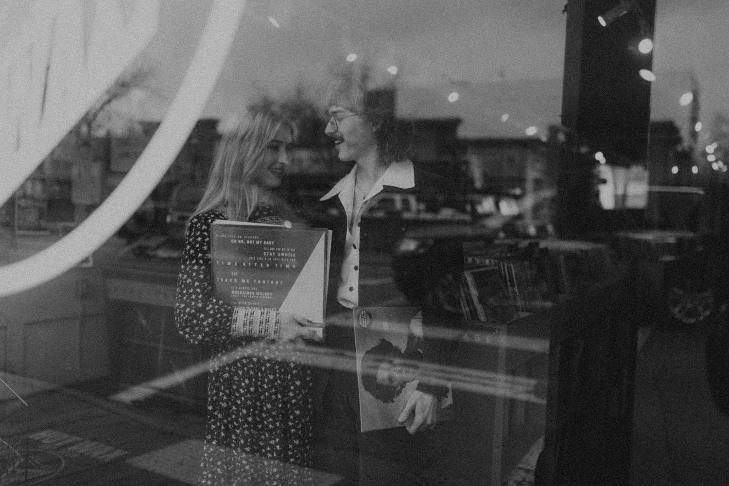 Black and white reflection in storefront window shows a figure with a shopping cart and city lights in background.