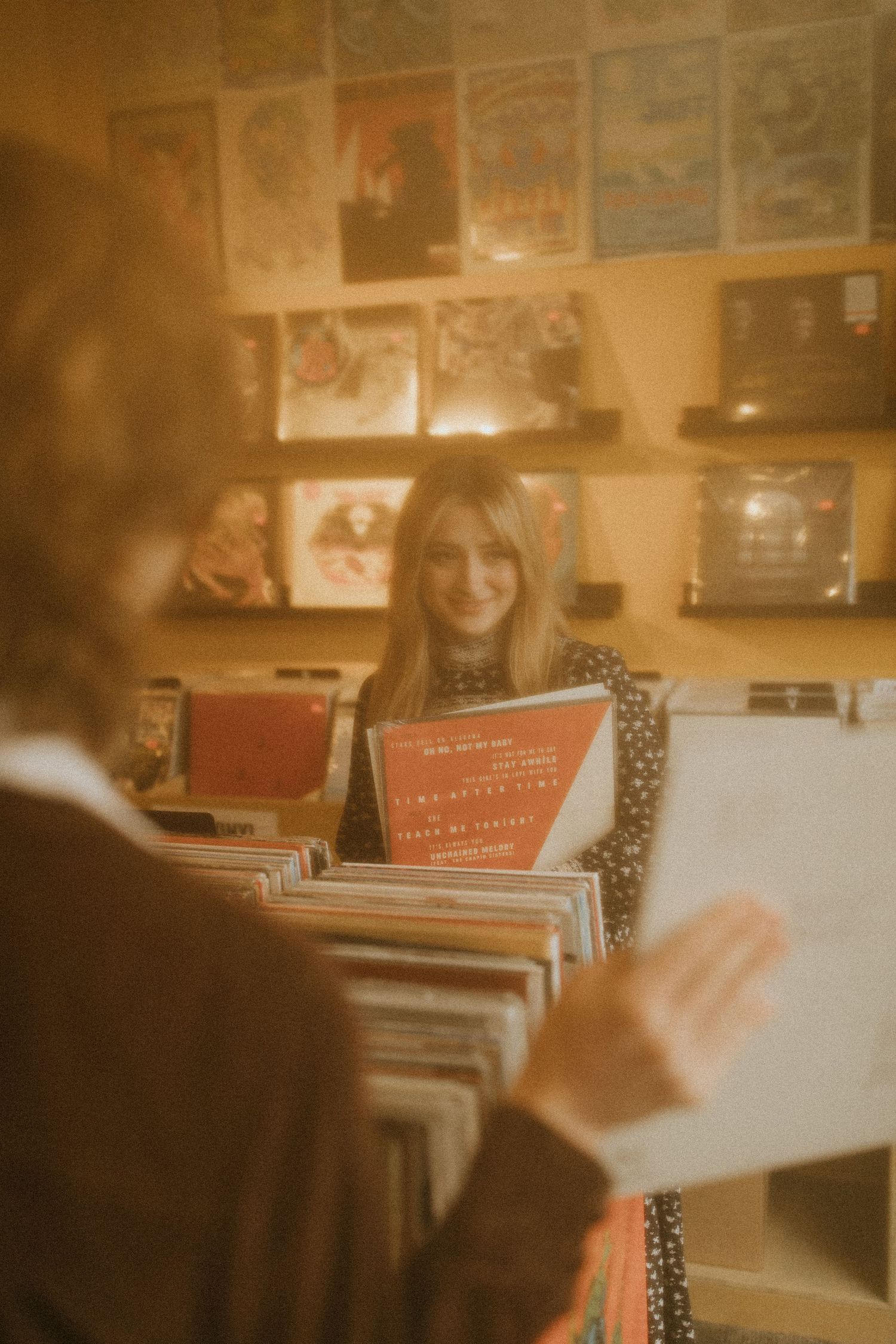 A warm vintage-tinted photograph shows someone browsing through vinyl records in a music store with posters on the wall.