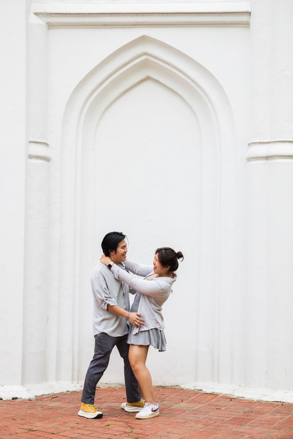 A romantic couple pose against a white Gothic archway with brick flooring.