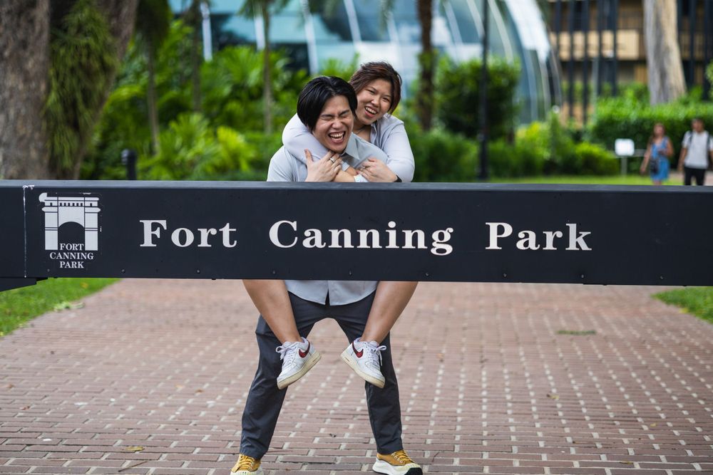 A playful moment captured at Fort Canning Park in Singapore with a couple posing against a black entrance sign.