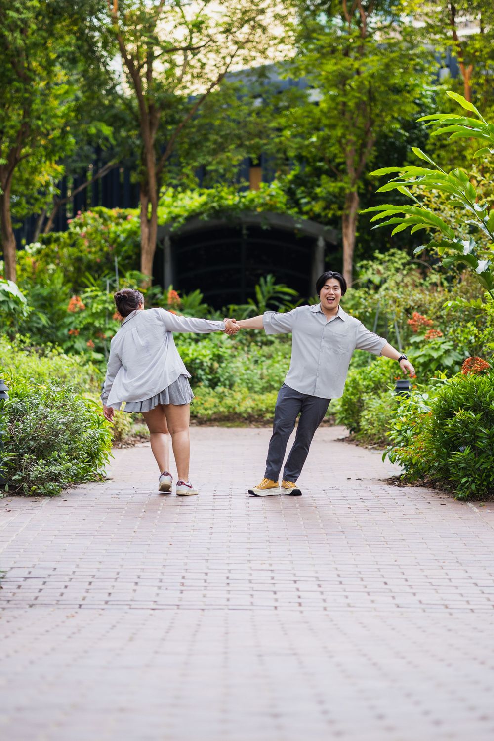 Couple holds hands while dancing along garden pathway surrounded by greenery.