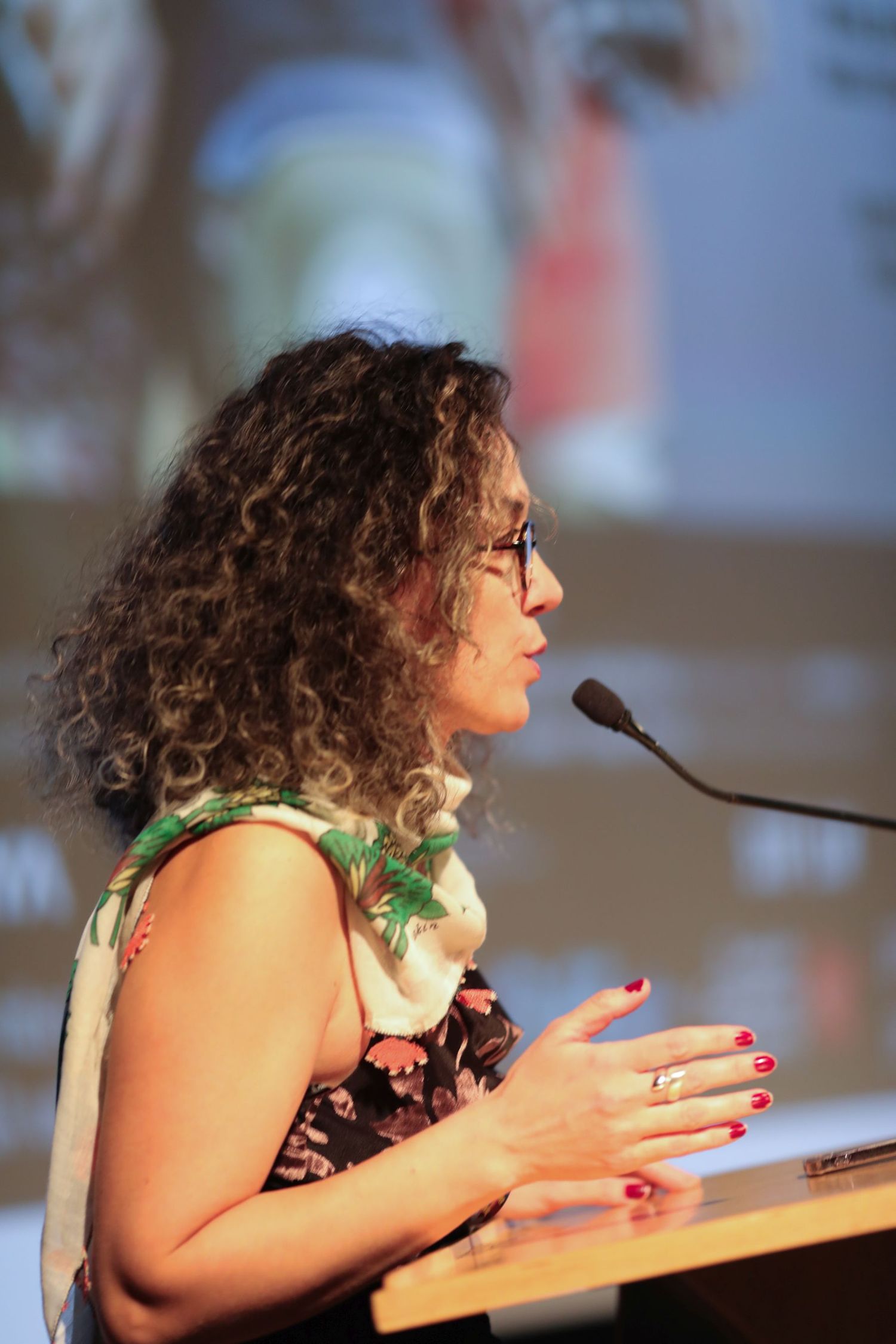 Speaker at a podium gestures while giving a presentation, wearing glasses and a floral accessory.