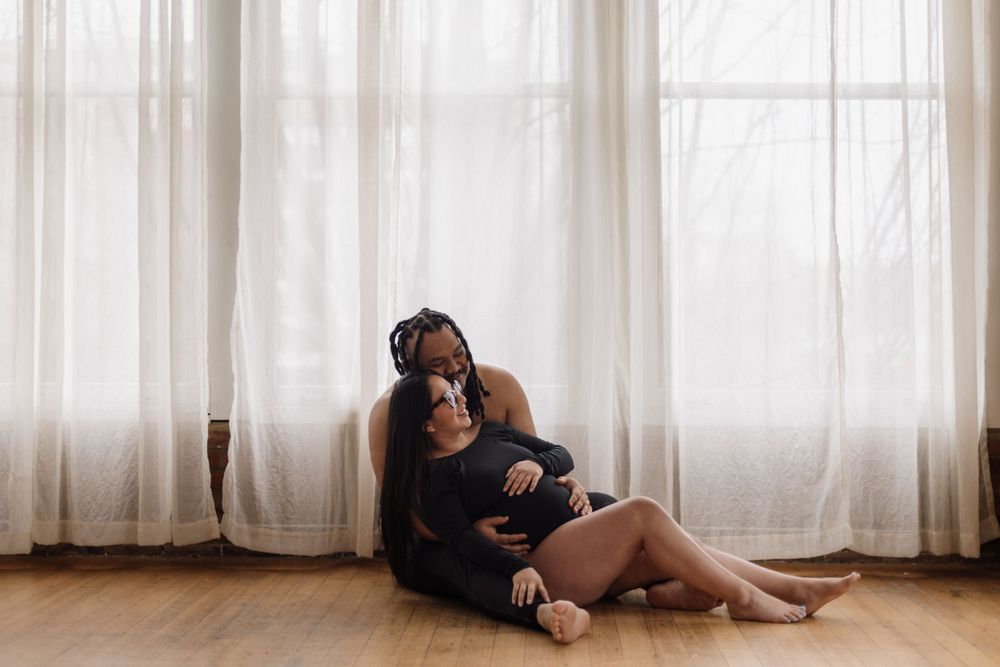 A relaxed portrait of someone sitting on hardwood flooring against sheer curtains in gentle window light.