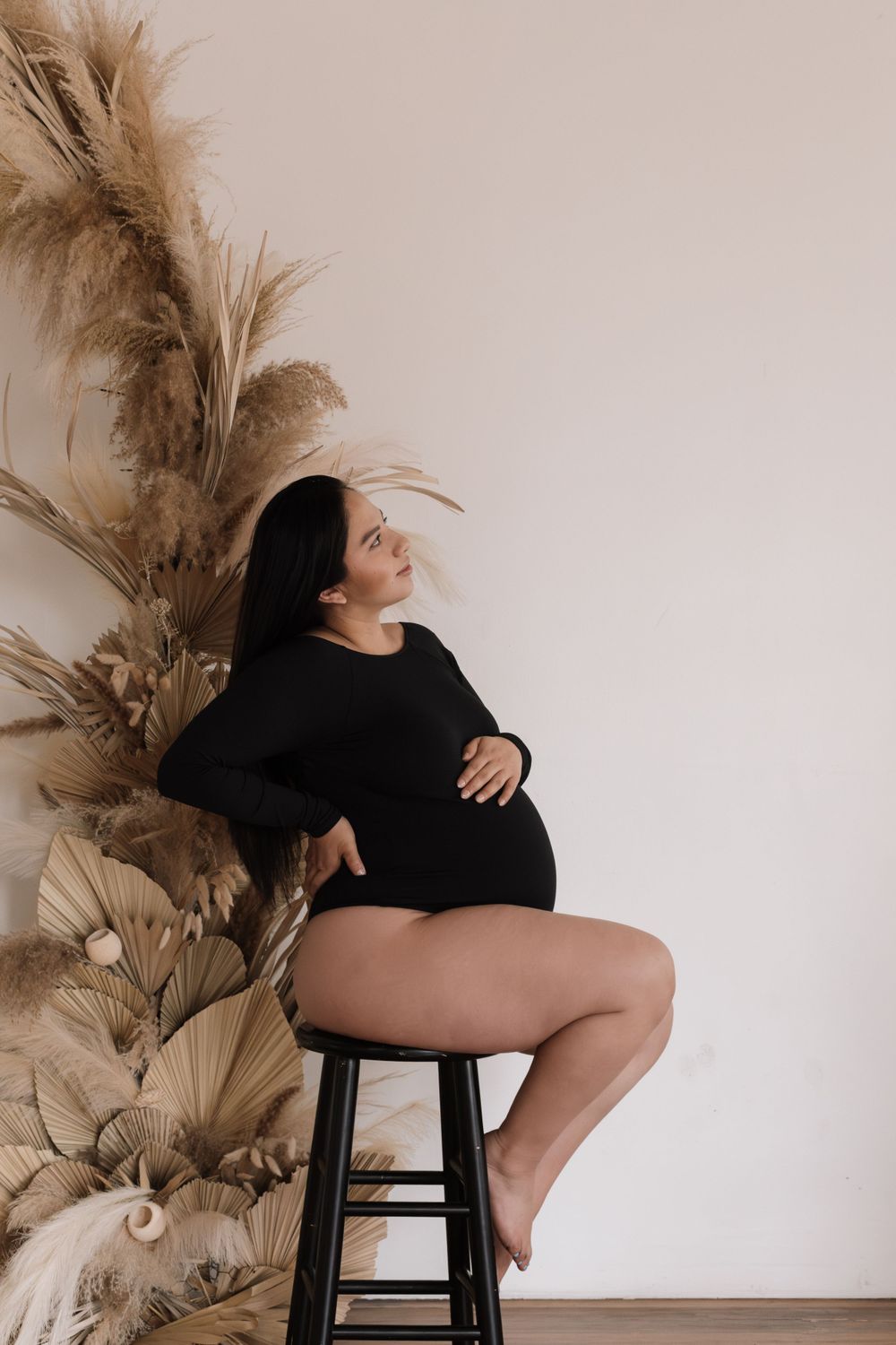 Artistic maternity photo with pampas grass decor featuring a woman in black sitting on a stool against neutral background.
