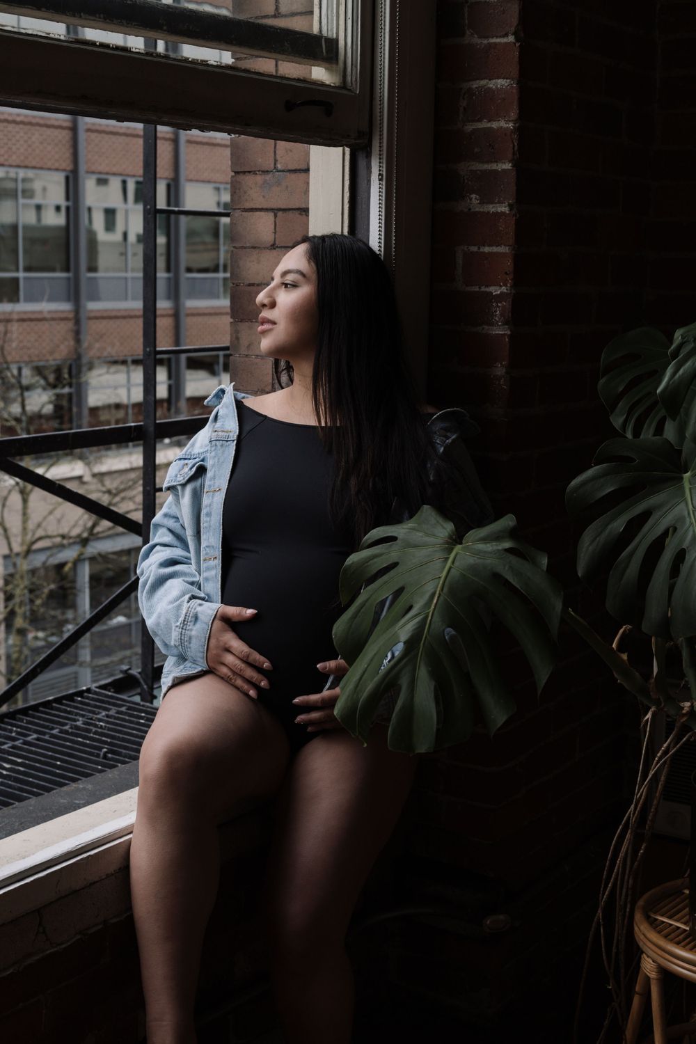 A person in a denim jacket and black bodysuit poses near a window with a monstera plant in moody lighting.
