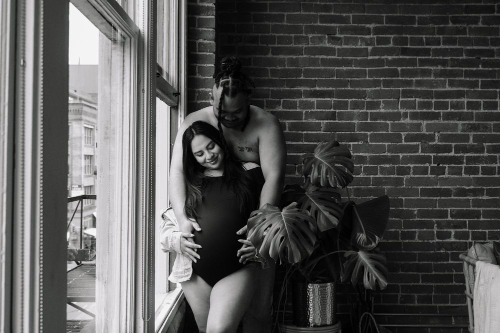 A couple shares an intimate black and white moment by a window with exposed brick walls and a potted plant.