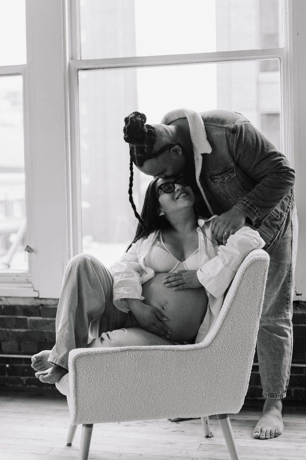 Intimate black and white maternity photo showing tender moment between couple in front of bright window.