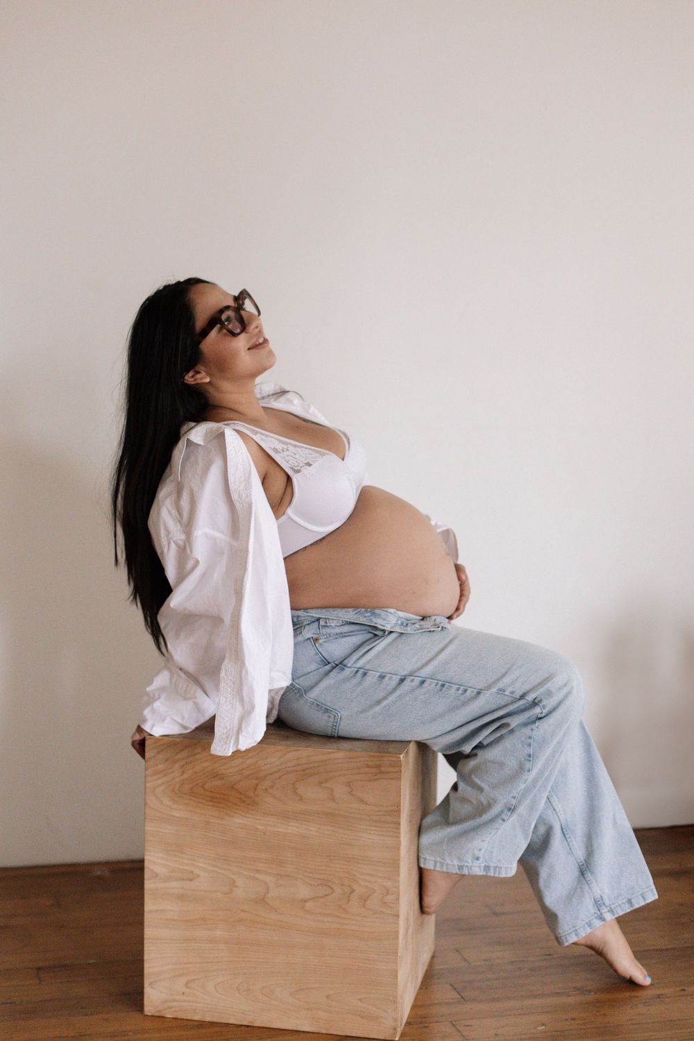 Side view of an expectant parent wearing loose jeans and white shirt, sitting casually on a wooden box indoors.