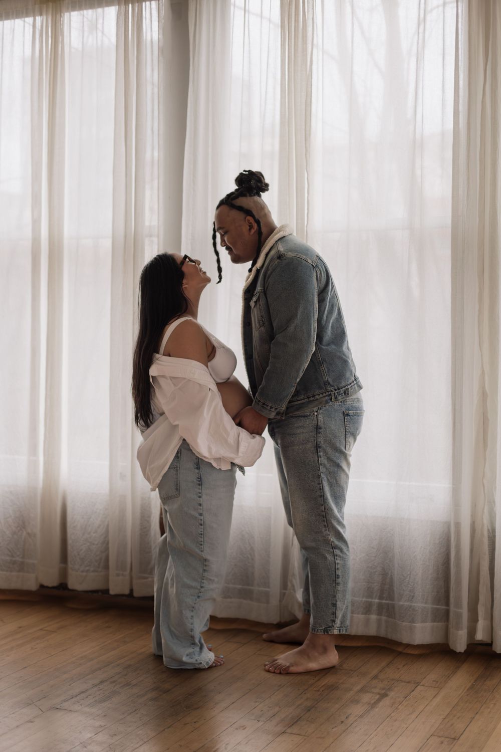 A tender moment between two people in jeans and casual wear standing by sheer curtains in natural light.