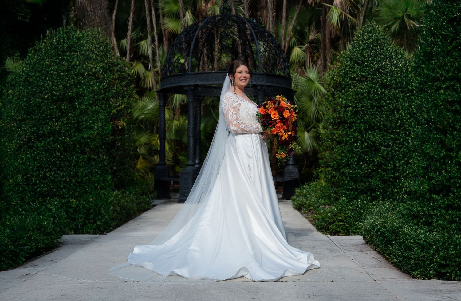 A bride in an elegant white long-sleeved wedding gown stands on a garden path holding a vibrant orange bouquet.