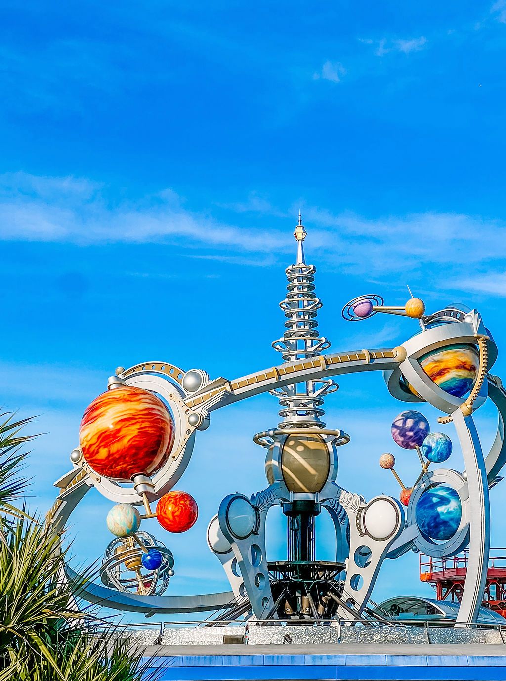 Colorful planetary-themed sculpture with orbiting spheres against bright blue sky at Tomorrowland in Disney theme park.