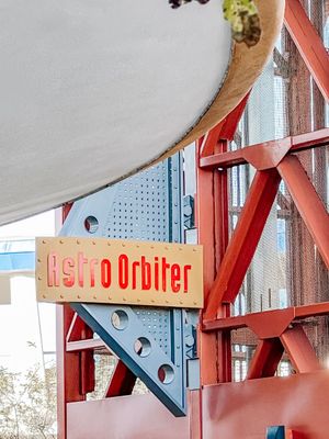 A red and blue retro-styled sign for 'Astro Orbiter' attraction mounted on industrial metal framework.