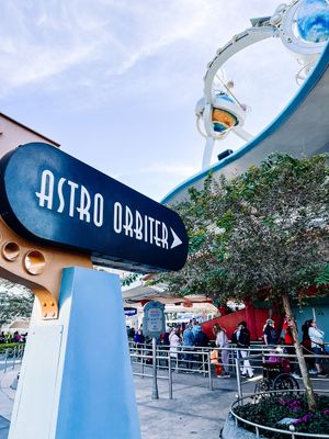 Retro 'Astro Orbiter' attraction sign with blue accents against a cloudy sky at a theme park entrance.