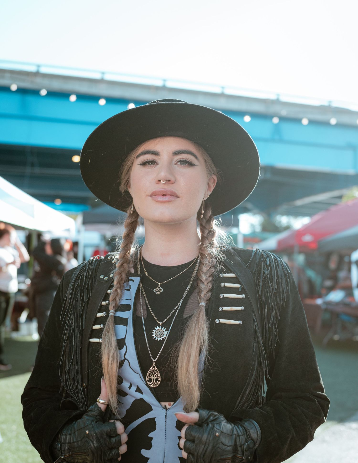 Person in western style outfit with wide brim hat and fringe jacket at an outdoor market.