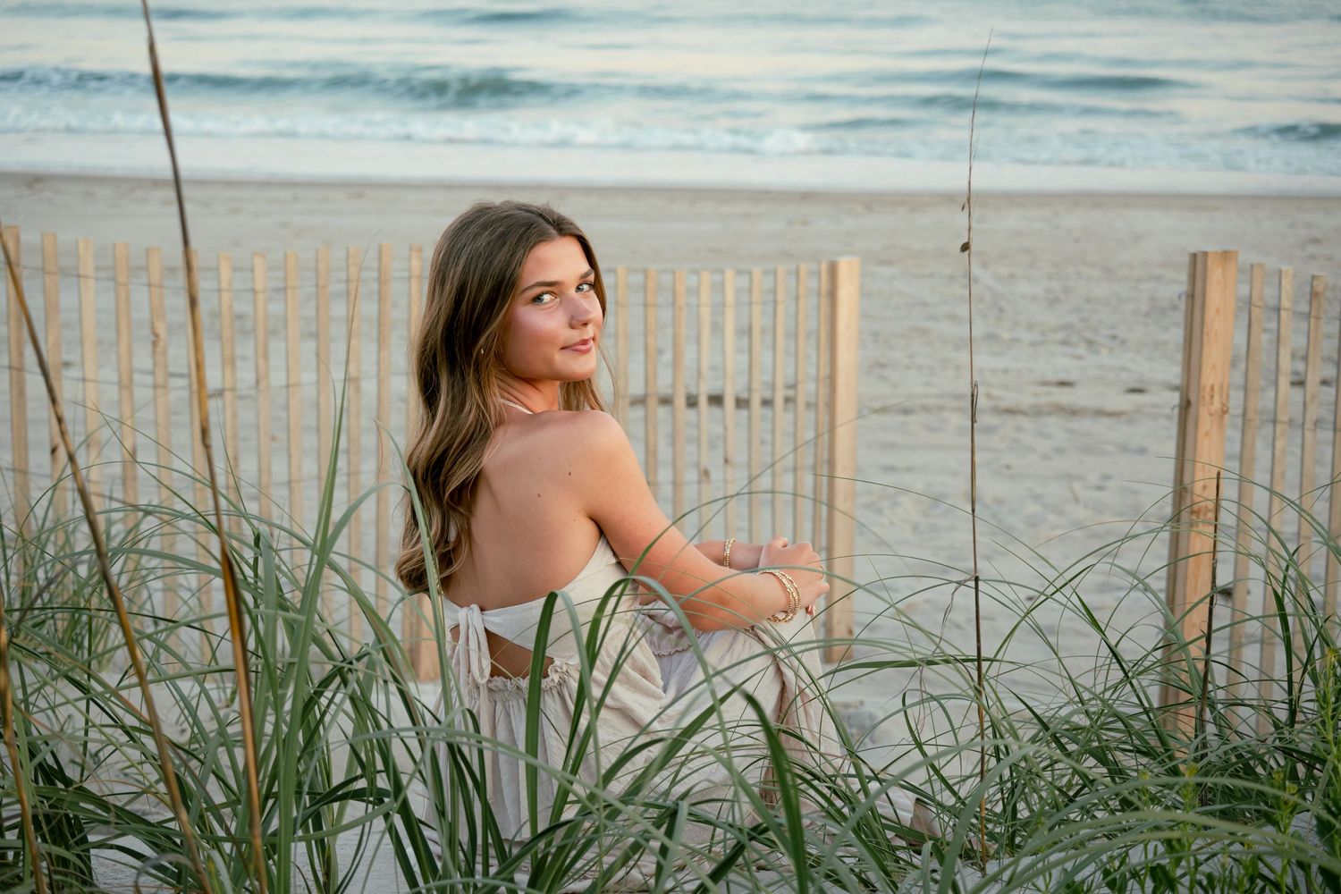Woman in white dress sitting among beach grass at sunset.