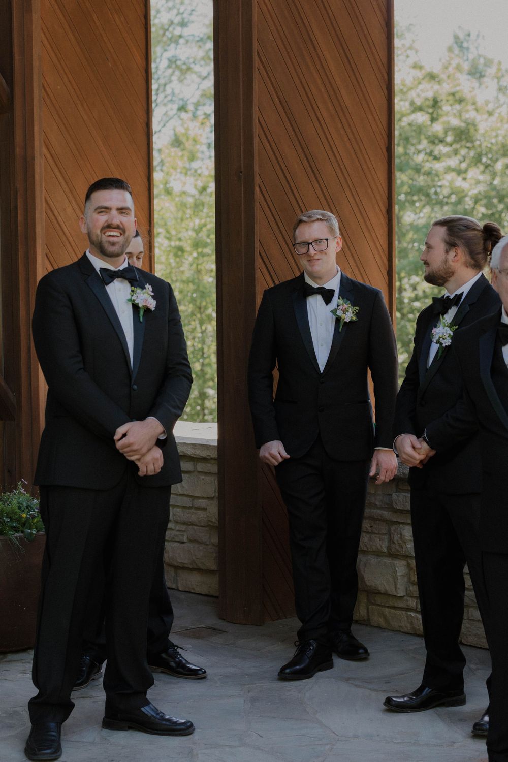 Wedding party members in black tuxedos standing on stone patio under wooden beams.