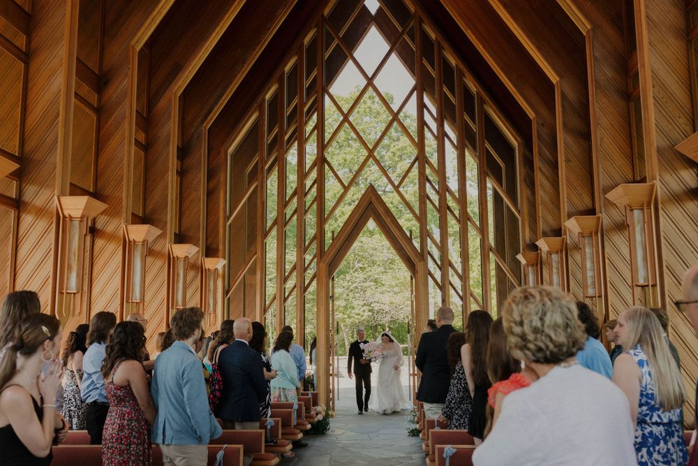 Wedding ceremony in progress inside wooden chapel with guests seated in pews.