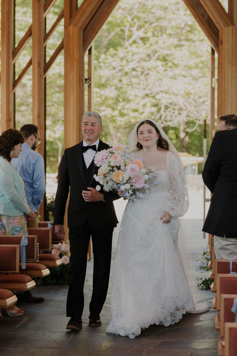 A couple in wedding attire stands in a rustic wooden pavilion surrounded by guests and floral decorations.