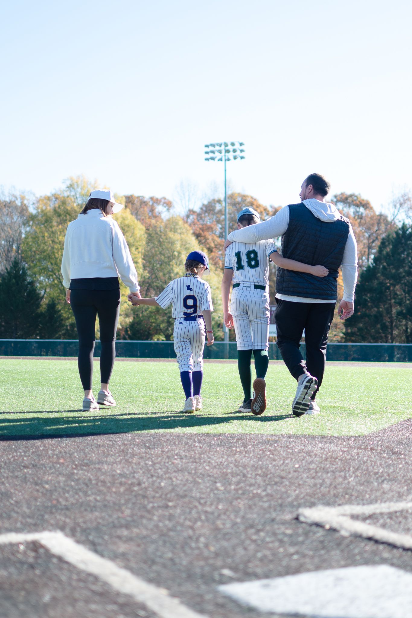 Baseball players walk on field near home plate with stadium lights and autumn trees in background during practice.