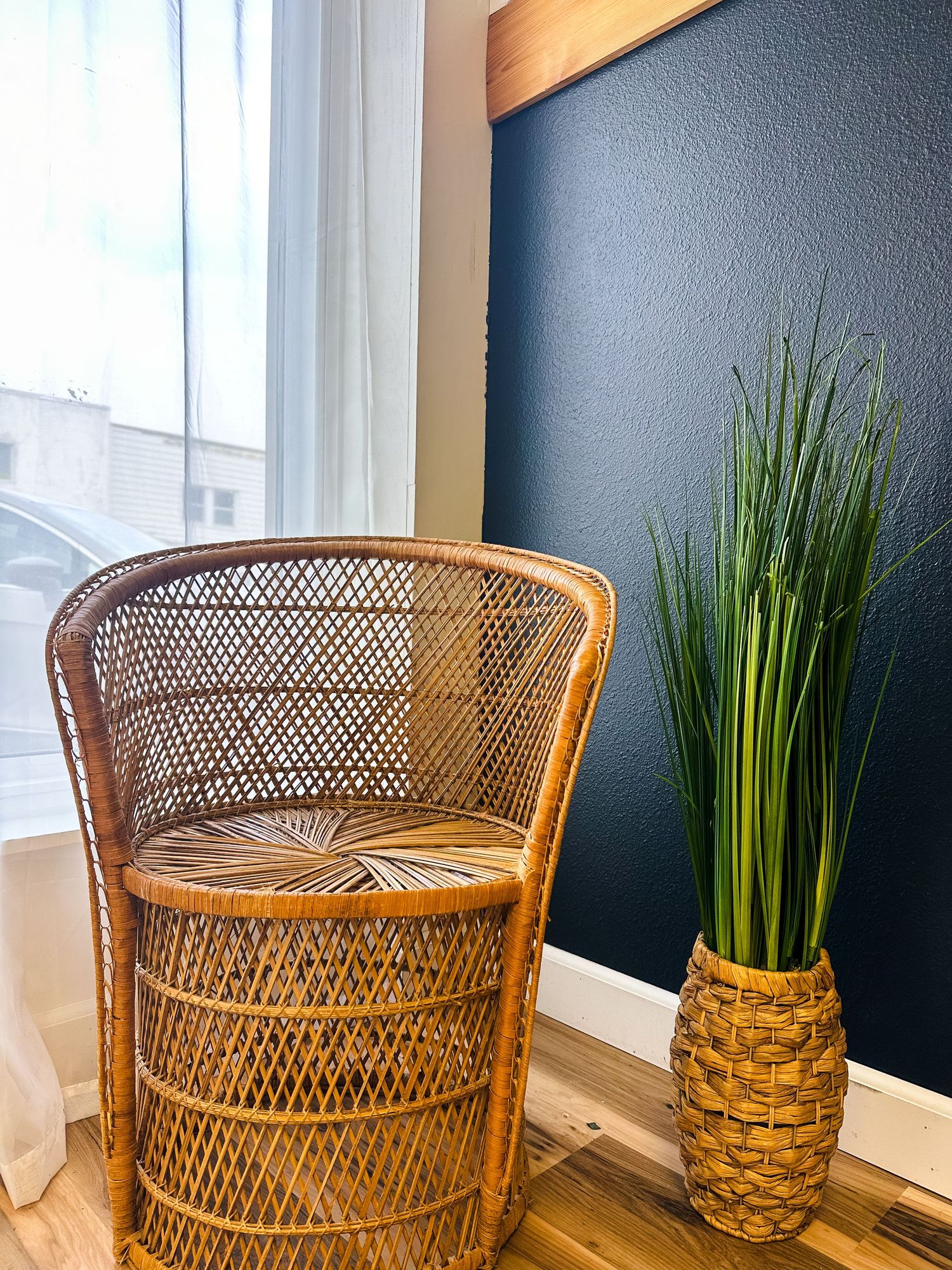 Vintage wicker peacock chair sits elegantly against navy wall with tall decorative grass plant in woven pineapple vase.