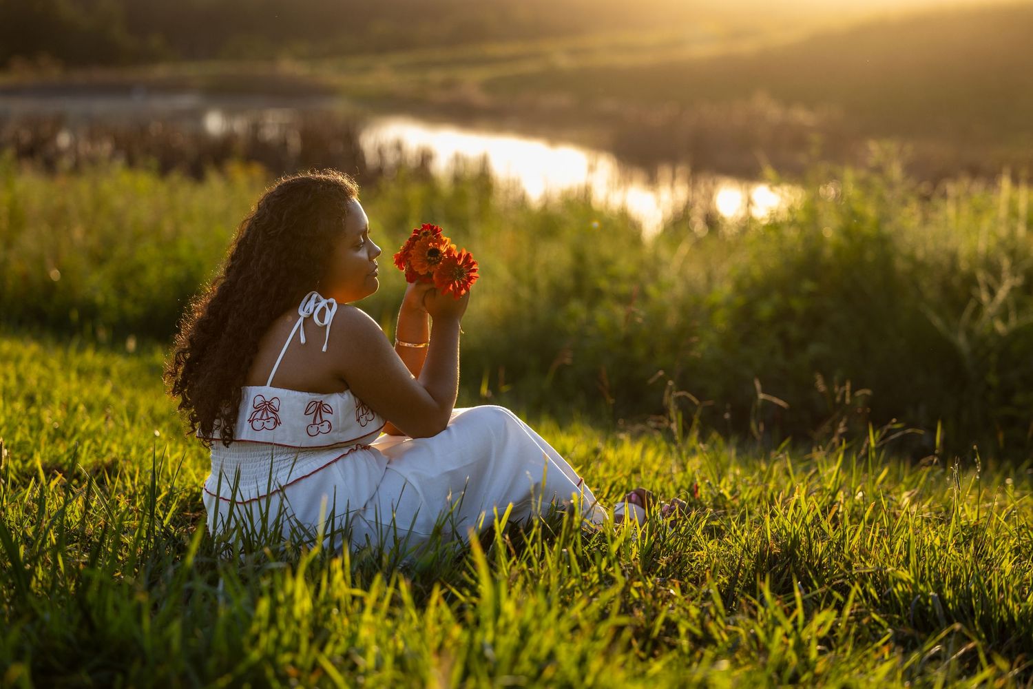 A person in a floral dress poses outdoors in soft golden sunlight while leaning on a surface.