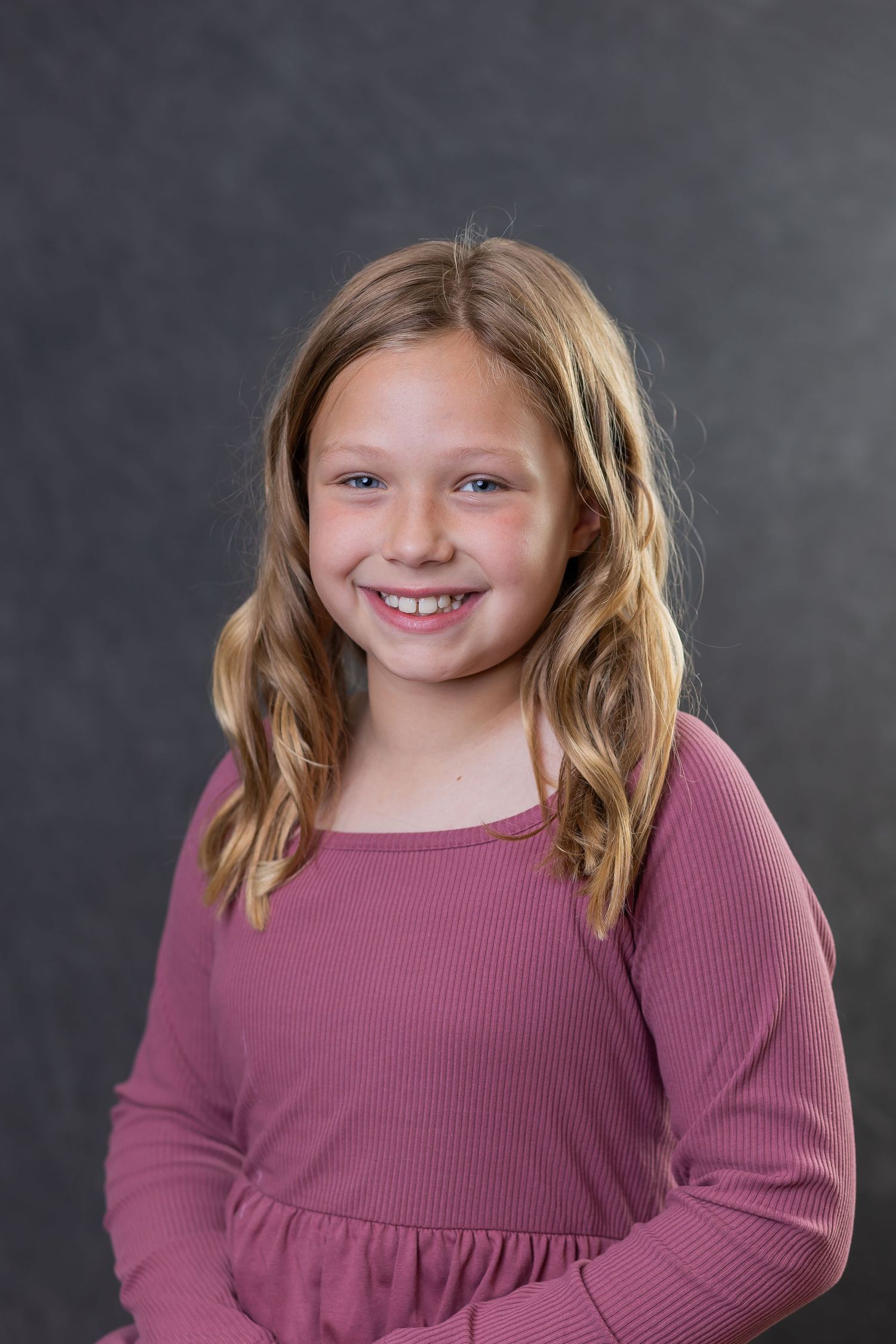 Young student in a mauve ribbed sweater poses against a gray backdrop for a school portrait.