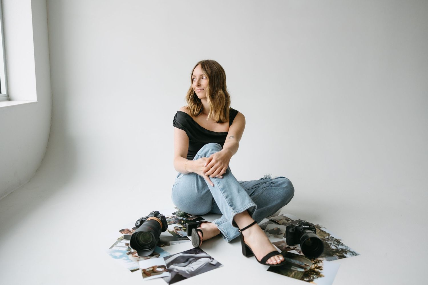 Woman in black top and jeans sits on floor surrounded by camera equipment near window.