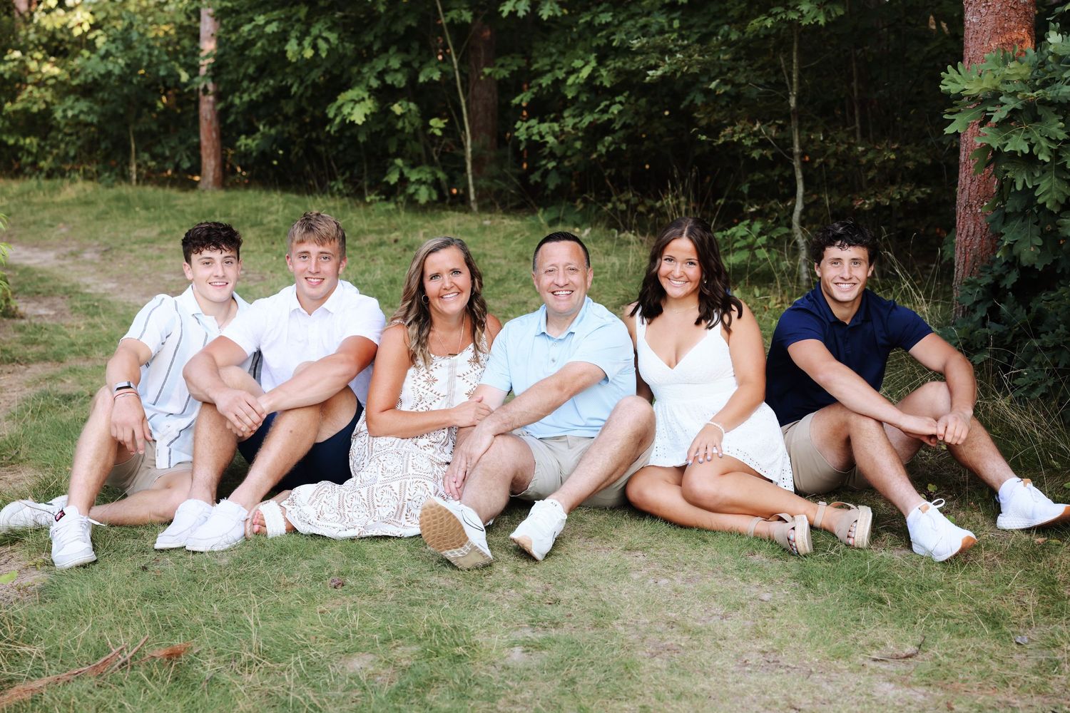 A group sits casually together on grass wearing coordinated white and light colored outfits on a summer day.