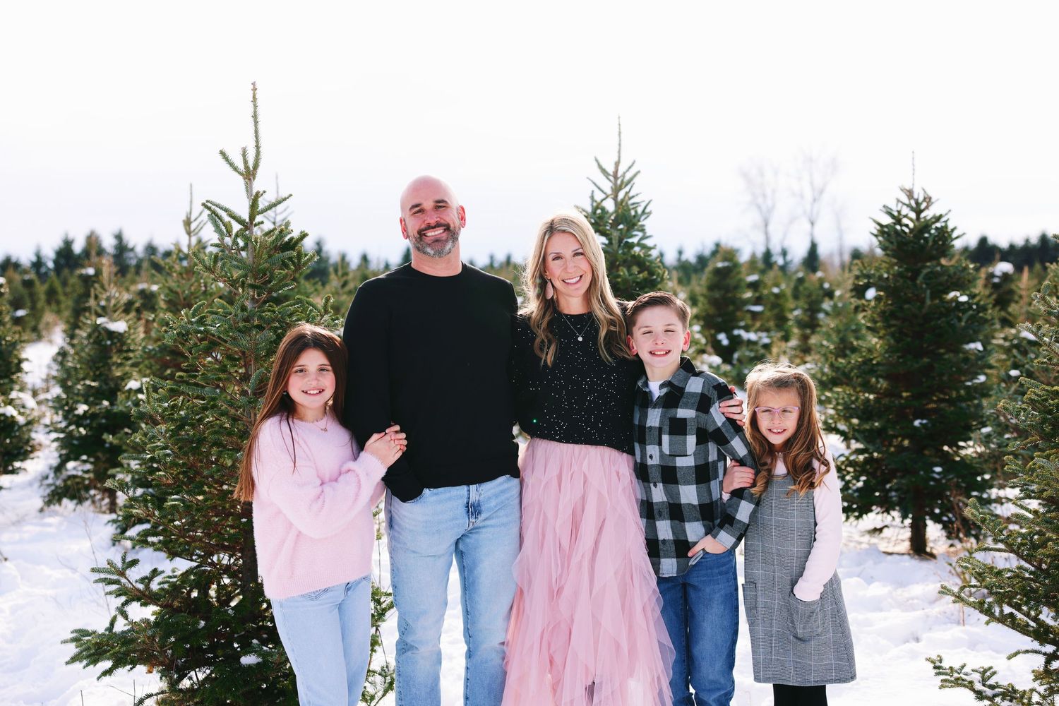 A family poses together in a snowy evergreen forest setting wearing coordinated winter outfits.