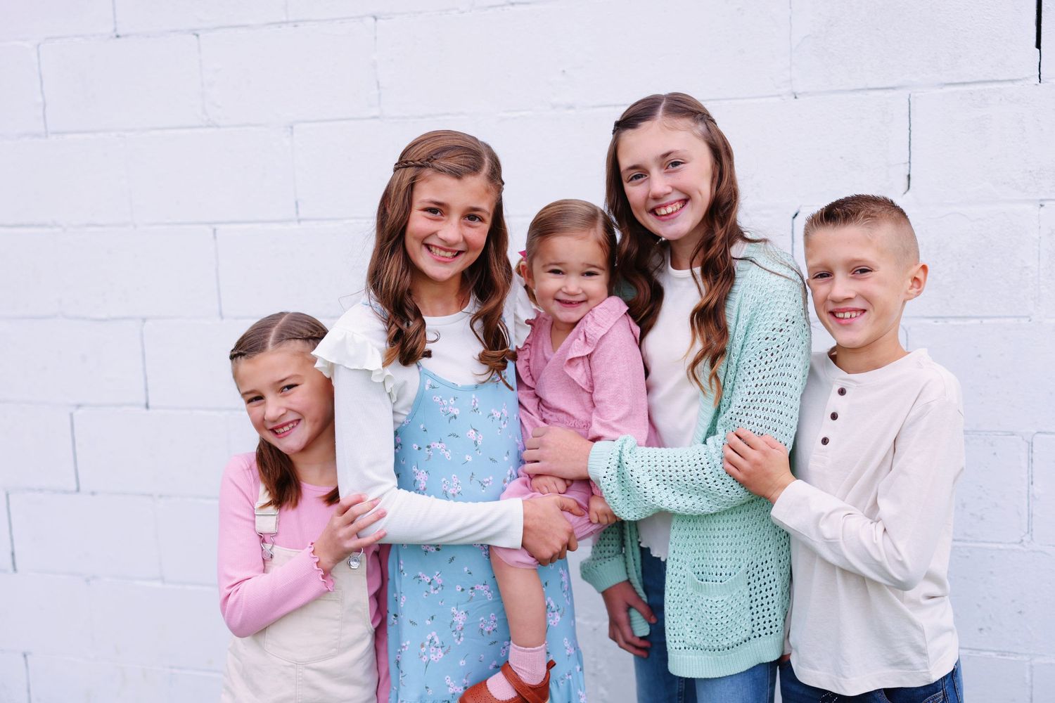 A group of children wearing pastel colored clothing stand together against a white brick wall.