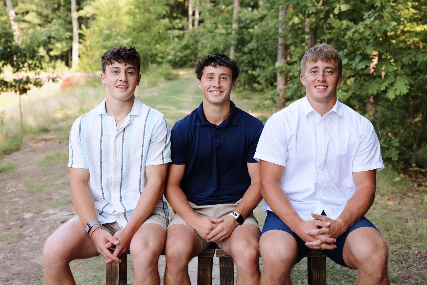 Three young men in casual summer attire sit together on a wooden bench along a wooded path on a sunny day.