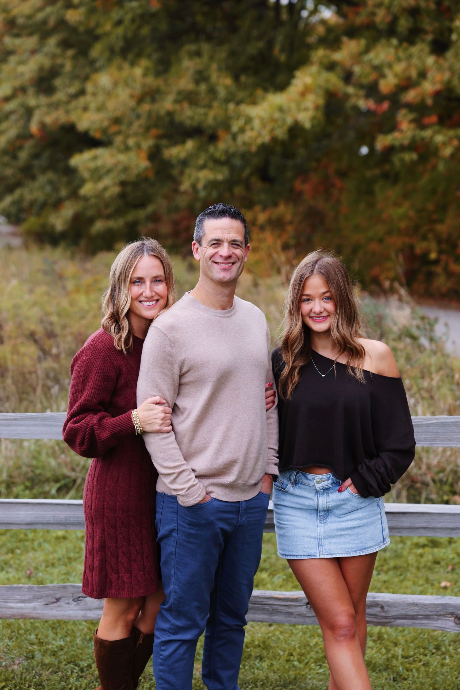 Three friends pose together outdoors in casual fall attire with autumn foliage in the background.