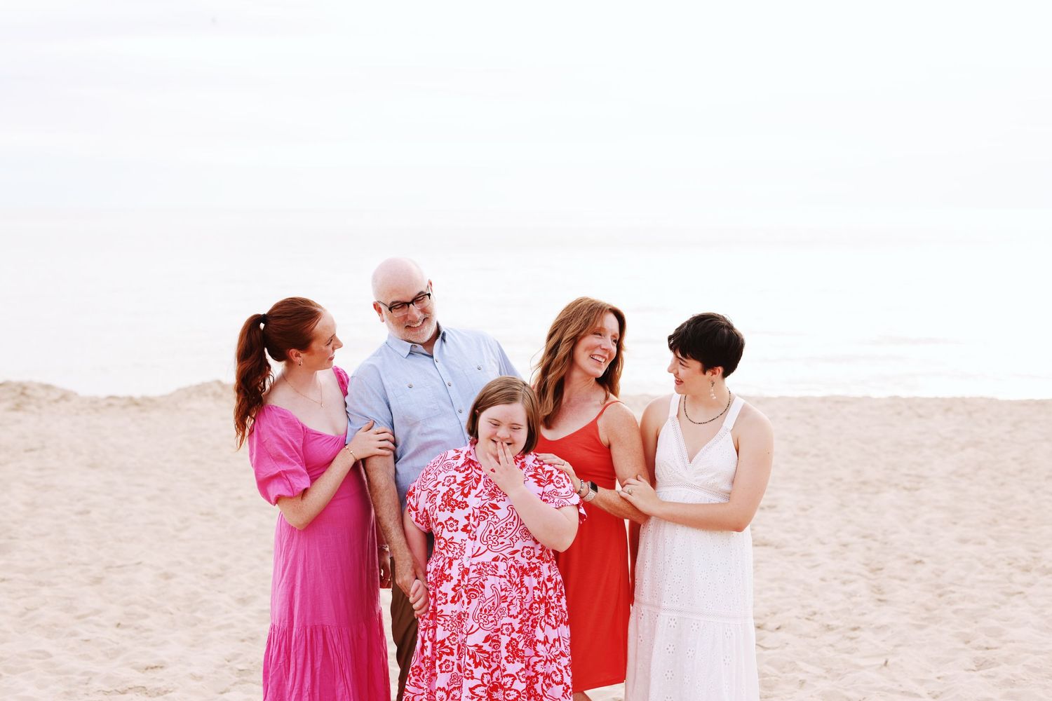 Multi-generational family wearing vibrant summer dresses stands together smiling on a bright sandy beach.
