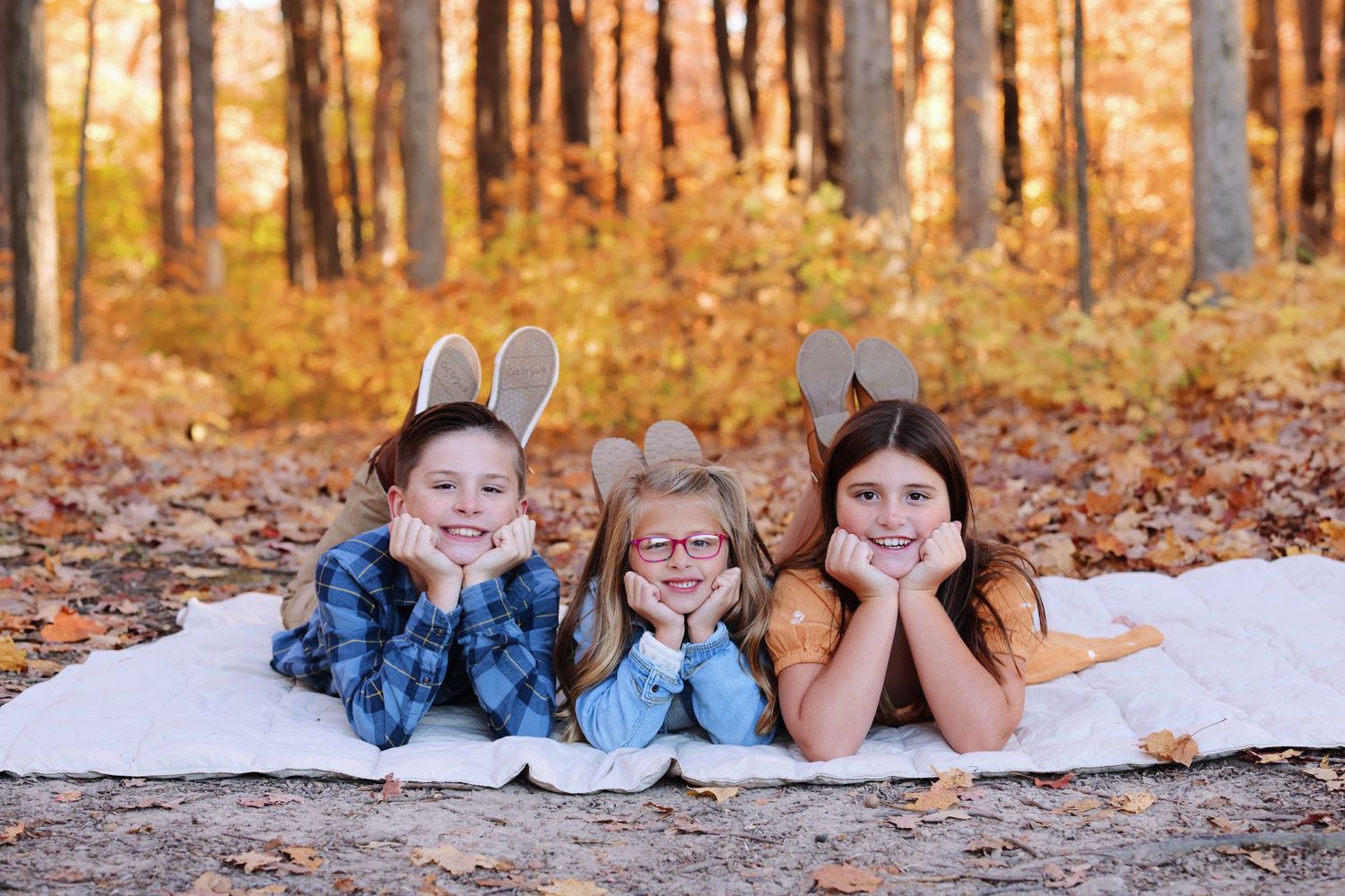 Three children lying on a white blanket in autumn forest with golden leaves in the background.