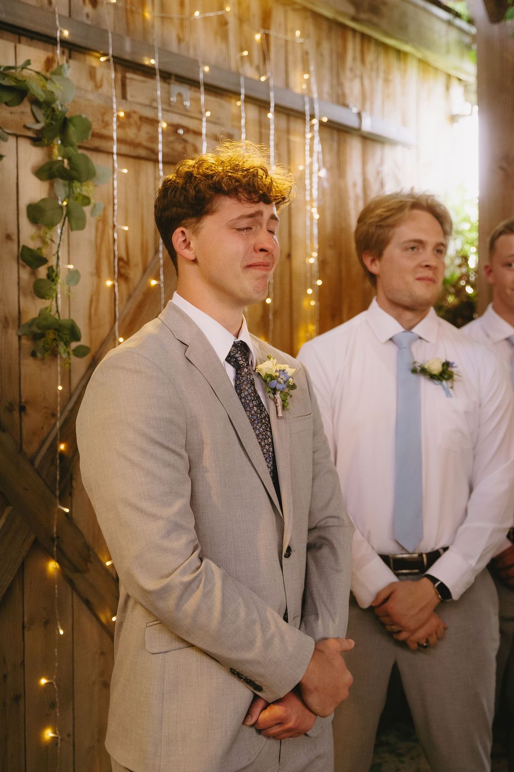 Groomsmen in light gray suits and ties stand in line during a wedding ceremony with twinkle lights in background.