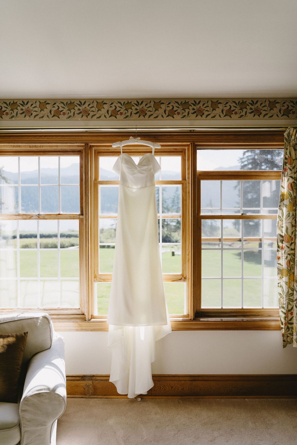An elegant white wedding dress hangs in front of large windows with a scenic mountain view.