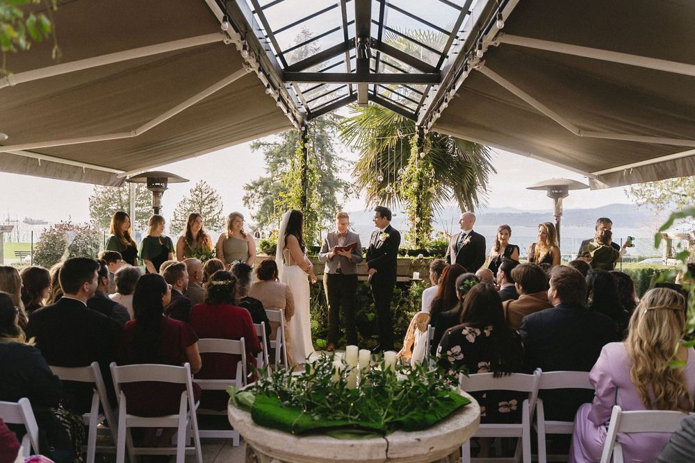 Wedding ceremony guests seated in white chairs under a conservatory-style roof with natural lighting.