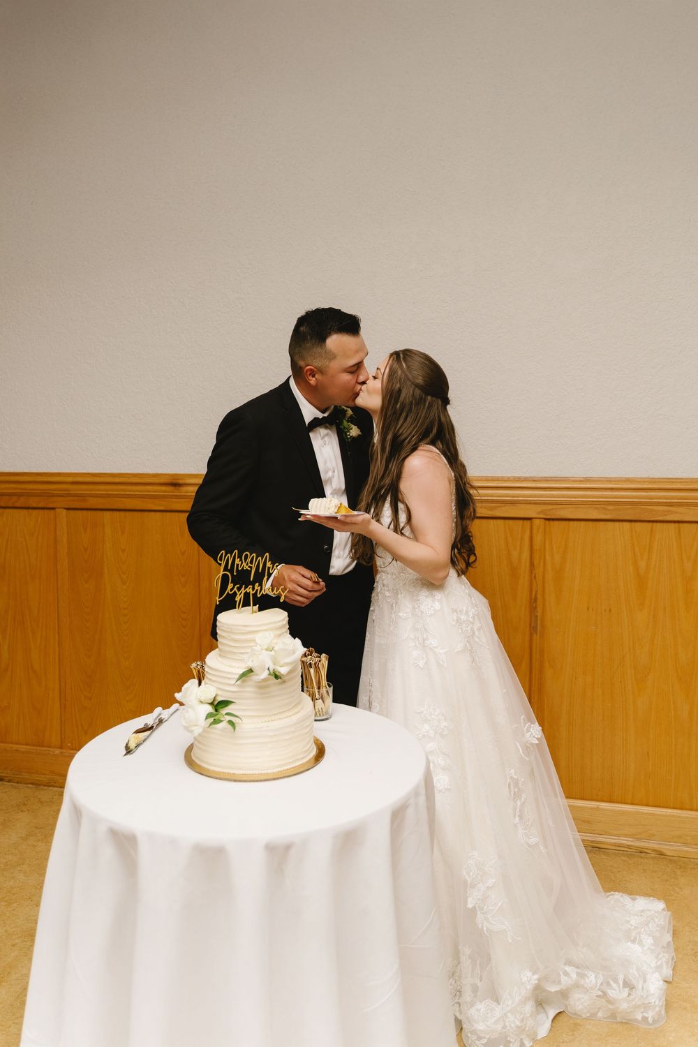 A wedding couple shares a romantic kiss while cutting their elegant white tiered cake at their reception.