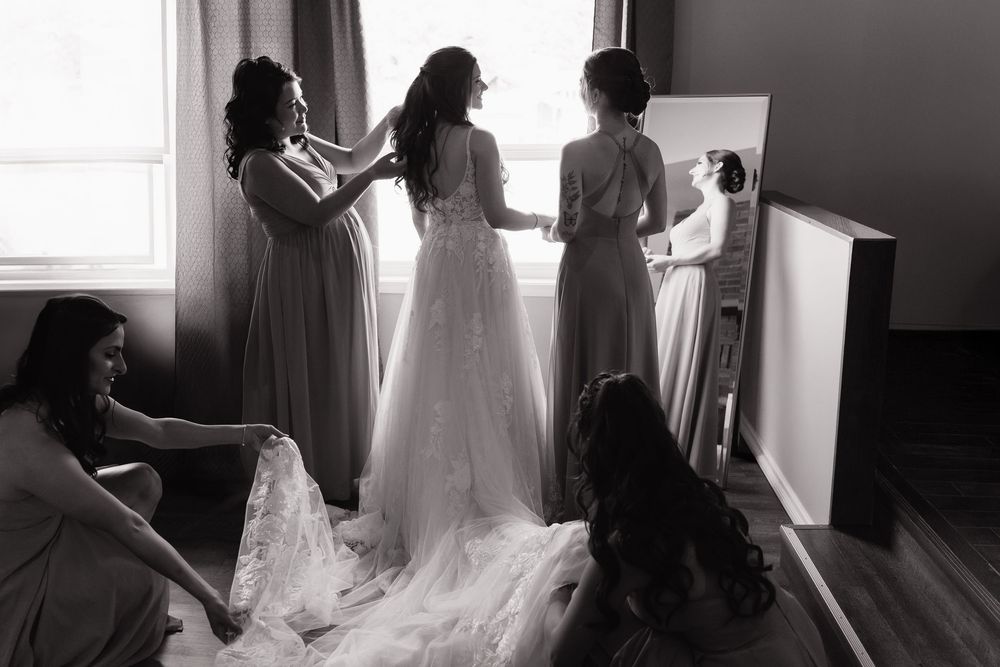 A bride in a flowing white wedding dress gets ready with her bridesmaids in a sunlit room.